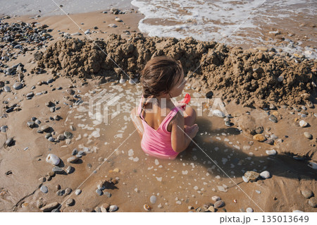 Little girl in a pink swimsuit playing actively with a red toy on the wet sand, building a moat and fort on a sunny pebble beach during a summer vacation Little girl in a pink swimsuit playing actively with a red toy on the wet sand, building a moat and fort on a sunny pebble beach during a summer vacation 135013649