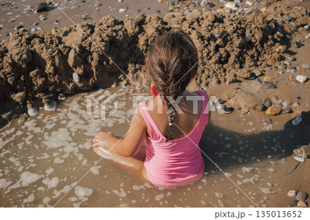 Little girl in a pink swimsuit playing actively with a red toy on the wet sand, building a moat and fort on a sunny pebble beach during a summer vacation Little girl in a pink swimsuit playing actively with a red toy on the wet sand, building a moat and fort on a sunny pebble beach during a summer vacation 135013652