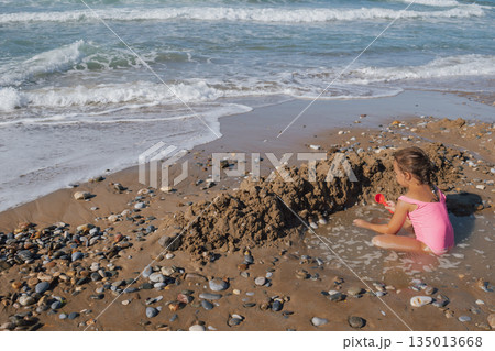 Happy Little Girl in Pink Swimsuit Building Sand Castle Moat on a Pebble Beach with Waves and Sea Foam in Summer Sunlight 135013668
