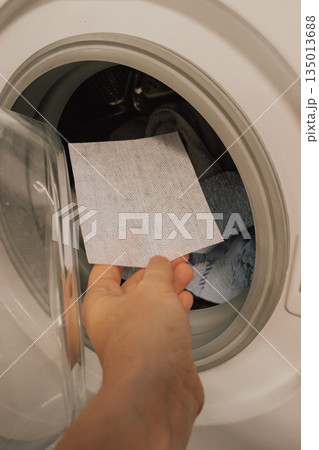 Hand placing a color catching laundry sheet into the washing machine drum with clothes. Preventing dye transfer and protecting garments from running colors during the wash cycle 135013688