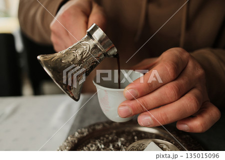 Close up of Bosnian coffee being poured into a small cup from a traditional cezve. Concept of detail, tradition and authentic Balkan ritual Close up of Bosnian coffee being poured into a small cup from a traditional cezve. Concept of detail, tradition and authentic Balkan ritual 135015096