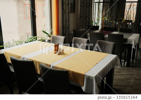 Empty restaurant dining area with neatly arranged tables and soft daylight from large windows. Hospitality and interior concept representing calm atmosphere before guests arrive Empty restaurant dining area with neatly arranged tables and soft daylight from large windows. Hospitality and interior concept representing calm atmosphere before guests arrive 135015164