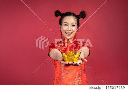 Portrait of smiling woman holding gold yuanbao toward camera on red background 135017489