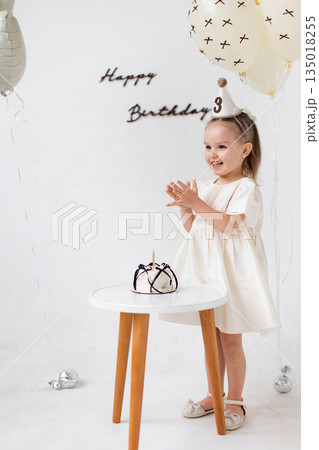 A young girl claps her hands with joy in front of a birthday cake, celebrating her third birthday with balloons and decorations. A young girl claps her hands with joy in front of a birthday cake, celebrating her third birthday with balloons and decorations. 135018255