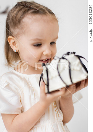 A young girl smiles while holding a small, decorative gift. She is wearing a white dress and has heart-shaped earrings. A young girl smiles while holding a small, decorative gift. She is wearing a white dress and has heart-shaped earrings. 135018284