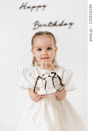 A young girl smiles while holding a birthday cake, with 'Happy Birthday' written on the wall behind her. 135018289