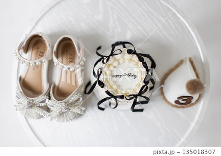 A pair of white shoes, a birthday cake, and a party hat are arranged on a white surface. 135018307