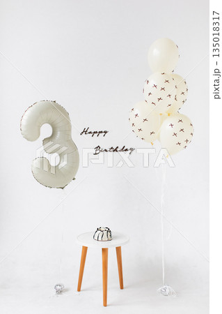 A minimalist birthday scene featuring a number three balloon, balloons, a small cake, and a 'Happy Birthday' message against a white backdrop. 135018317