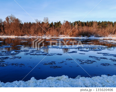 Frozen River with Floating Ice in Winter Landscape. Calm winter river partially covered with floating ice chunks reflecting trees and sky on cold sunny day. Frozen River with Floating Ice in Winter Landscape. Calm winter river partially covered with floating ice chunks reflecting trees and sky on cold sunny day. 135020406