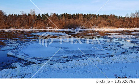 Winter River with Drifting Ice Floes at Sunset. Serene winter river scene with numerous floating ice chunks on water surface reflecting warm sunset light against forested shore. Winter River with Drifting Ice Floes at Sunset. Serene winter river scene with numerous floating ice chunks on water surface reflecting warm sunset light against forested shore. 135020768