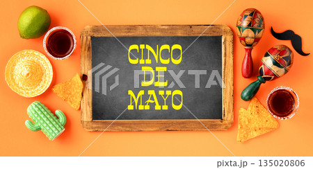 A vibrant Cinco de Mayo-inspired flat lay featuring vintage chalk board, maracas, tequila shots, lime, nacho chips on a bright orange backdrop 135020806