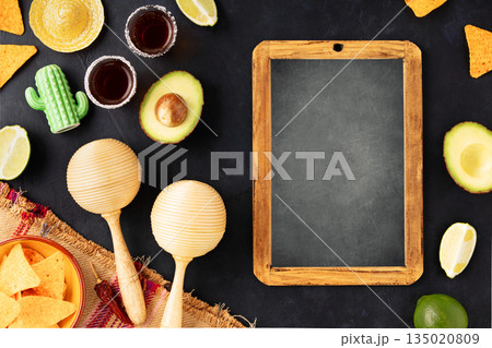 Cinco de Mayo-inspired flat lay featuring vintage chalk board, maracas, tequila shots, lime, nacho chips on a dark vintage background 135020809