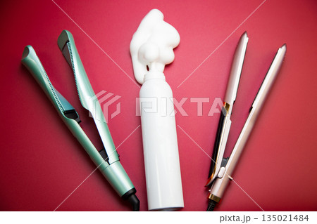 In the center are a white bottle of styling foam, around is a straightener and a curling iron to create hair curls on a pink background. The concept of hair care, creating a hairstyle and hair volume 135021484
