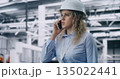 Female Engineer Wearing Hard Hat Walks Across Production Workshop and Speaks on Mobile Phone 135022441