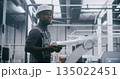 Factory Worker in Hard Hat Uses Handheld Device While Monitoring Robotic Equipment 135022451