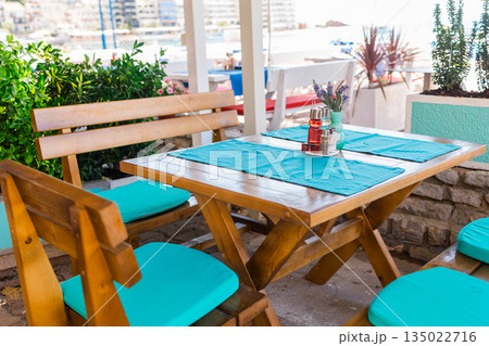 Wooden cafe table with turquoise cushions and table setting. Relaxed seaside dining, attention to detail, and welcoming atmosphere in coastal hospitality. 135022716