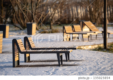 Empty wooden sunbeds for rest standing near river in city park covered in snow on sunny winter day. 135023854