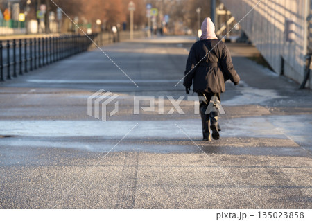 Woman walking on frozen asphalt sidewalk with technical salt and substances sprinkled over surface. 135023858