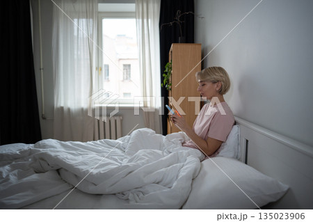 Lazy middle aged woman in bed scrolling online on smartphone, procrastination and phone addiction. 135023906