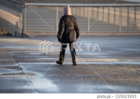 Woman walking on frozen asphalt sidewalk with technical salt and substances sprinkled over surface. 135023933
