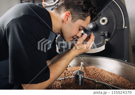 Side view of young worker smelling fresh roasted coffee beans above the machine drum 135024370