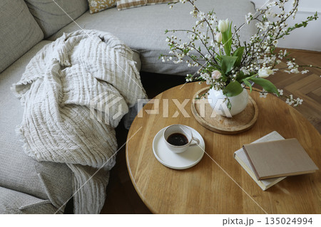 Cozy Scandinavian living room still life with coffee cup, books. Spring flowers, tulips and blooming cherry branches in vase on round wooden table. Elegant home interior lifestyle Easter morning scene 135024994