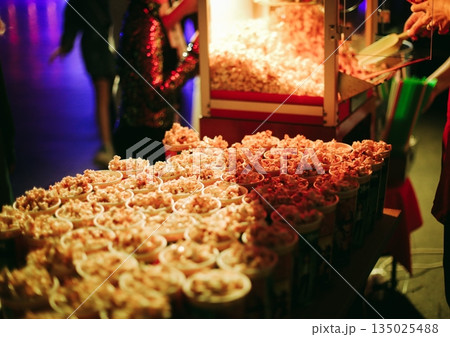 Popcorn cups stacked under neon lights, busy night market scene, vendor restocking containers, colorful stage lighting reflecting on snack display Popcorn cups stacked under neon lights, busy night market scene, vendor restocking containers, colorful stage lighting reflecting on snack display 135025488