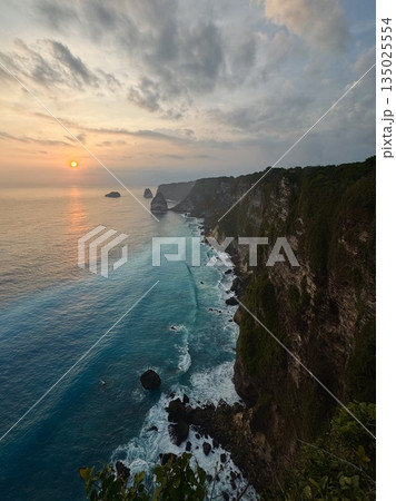 Nusa Penida, Indonesia, rocks, ocean, balisunset light spilling across towering coastal cliffs, golden glow on rock faces, soft cloudscape, silhouette of distant islets, calm sea reflecting warm 135025554