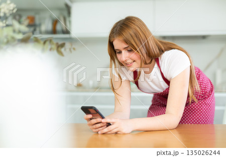 serious young girl stands in kitchen in red apron and holds smartphone in her hands and reads news serious young girl stands in kitchen in red apron and holds smartphone in her hands and reads news 135026244
