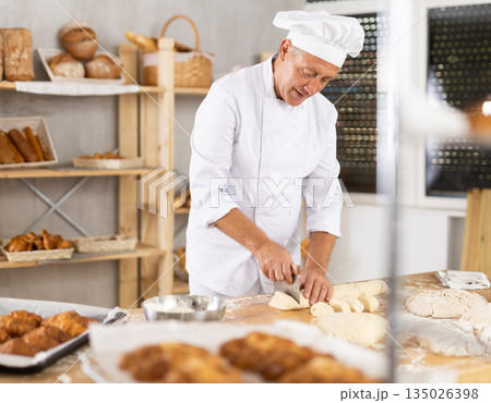Senior man works in bakery as baker, cuts dough into portions, forms pieces of dough to create buns. 135026398