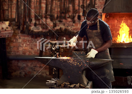 Blacksmith, worker and metal forge in workshop and manual industry working on hot steel with hammer, sparks or fire. Black man, welding or iron tools manufacturing or expert, trade and dark workspace Blacksmith, worker and metal forge in workshop and manual industry working on hot steel with hammer, sparks or fire. Black man, welding or iron tools manufacturing or expert, trade and dark workspace 135026593