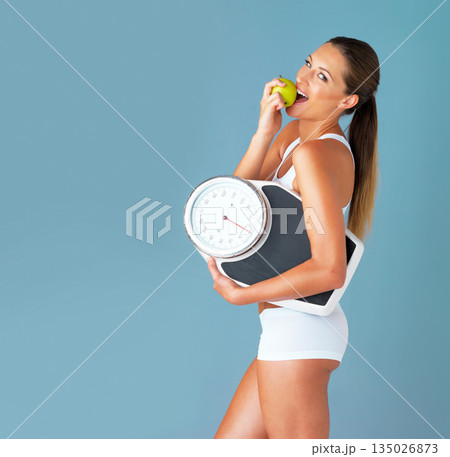 Woman is eating apple, fruit and scale with diet and health for weight loss in portrait isolated on blue background. Healthy food, female person and organic in studio with wellness and mockup space 135026873
