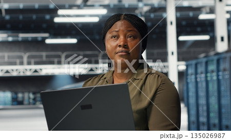 Technician in server room using laptop to upgrade rigs for increase in clients workload capacity. African american woman in data center configures equipment, improving processing speed, camera B Technician in server room using laptop to upgrade rigs for increase in clients workload capacity. African american woman in data center configures equipment, improving processing speed, camera B 135026987