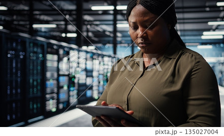 Data center software developer uses tablet to develop disaster recovery plans, minimizing downtime. IT worker in server hub enhancing data protection protocols using device, camera A close up 135027004