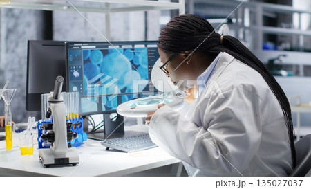 African american scientist uses magnifying glass to check Petri dish matter in biolab, laboratory experiment emphasizes microbiology and genetics research. Advancing medical research. 135027037