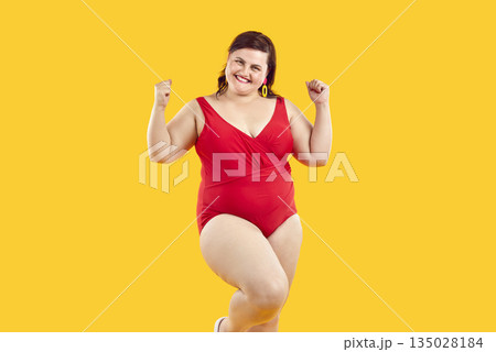 Happy joyful plus size female model in red swimsuit standing on yellow background Happy joyful plus size female model in red swimsuit standing on yellow background 135028184