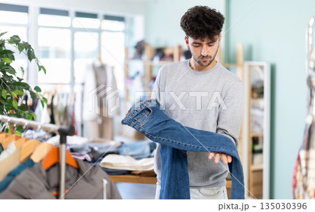 Interested guy inspecting pair of jeans in clothing store Interested guy inspecting pair of jeans in clothing store 135030396