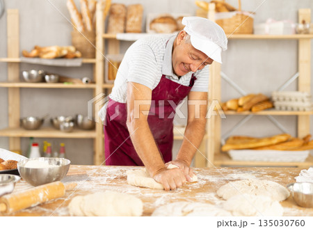 Elderly male baker kneading dough on table 135030760