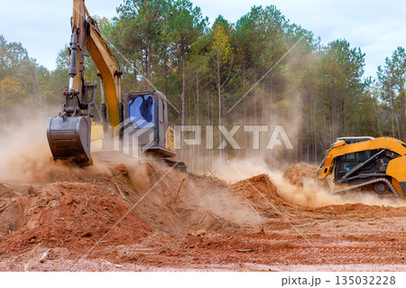 Construction excavator, equipment bulldozer operates in wooded area, removing dirt creating dust as trees are cleared for development. 135032228