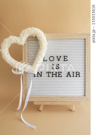 Letter board with text LOVE IS IN THE AIR Heart made of white lilac flowers on pastel beige background. Still life romantic composition of Valentines day holiday copy space for your text invitation 135033616