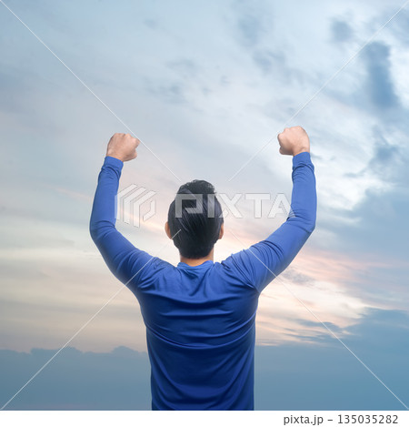 Rear view of man from the back with his arms raised up in front of a cloudscape at dusk, a feeling of freedom and achievement Rear view of man from the back with his arms raised up in front of a cloudscape at dusk, a feeling of freedom and achievement 135035282