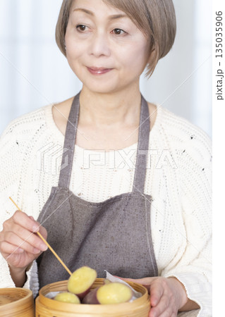 せいろでさつまいもを蒸して食べるシニア女性 135035906