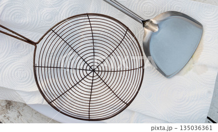Top view of wire mesh strainer and stainless steel spatula on white paper towel. Kitchen utensils for frying and draining oil concept. 135036361