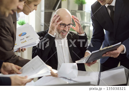 Stressed manager overwhelmed by paperwork as team pushes reports during deadline Stressed manager overwhelmed by paperwork as team pushes reports during deadline 135038321