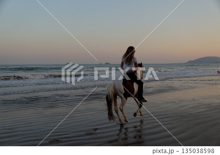 A young blonde girl rides a white horse along the seashore at sunset.Horse and sunset at sea. A young blonde girl rides a white horse along the seashore at sunset.Horse and sunset at sea. 135038598