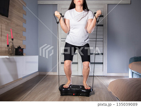 Brunette girl exercises with expanders at home on a vibration platform to reduce weight and cellulite. Exercise at home, lifestyle Brunette girl exercises with expanders at home on a vibration platform to reduce weight and cellulite. Exercise at home, lifestyle 135039089