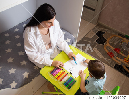 Caucasian woman and young boy drawing with colorful markers at green table in child room, creative play and learning activity, overhead perspective. Caucasian woman and young boy drawing with colorful markers at green table in child room, creative play and learning activity, overhead perspective. 135039097