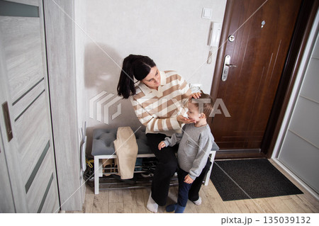 Woman caring for young child in home entryway, adjusting childs face before leaving. Family moment of connection in daily life. Woman caring for young child in home entryway, adjusting childs face before leaving. Family moment of connection in daily life. 135039132