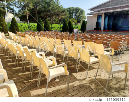 Empty chairs arranged in rows for outdoor event, concert venue setup with stage, trees in background, sunny day Empty chairs arranged in rows for outdoor event, concert venue setup with stage, trees in background, sunny day 135039312