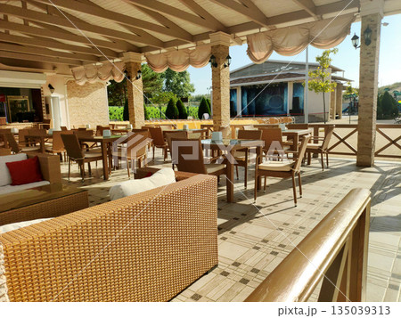 Outdoor cafe patio with wicker furniture under covered pergola, empty tables and chairs bathed in bright sunlight, open-air restaurant seating area awaiting guests Outdoor cafe patio with wicker furniture under covered pergola, empty tables and chairs bathed in bright sunlight, open-air restaurant seating area awaiting guests 135039313